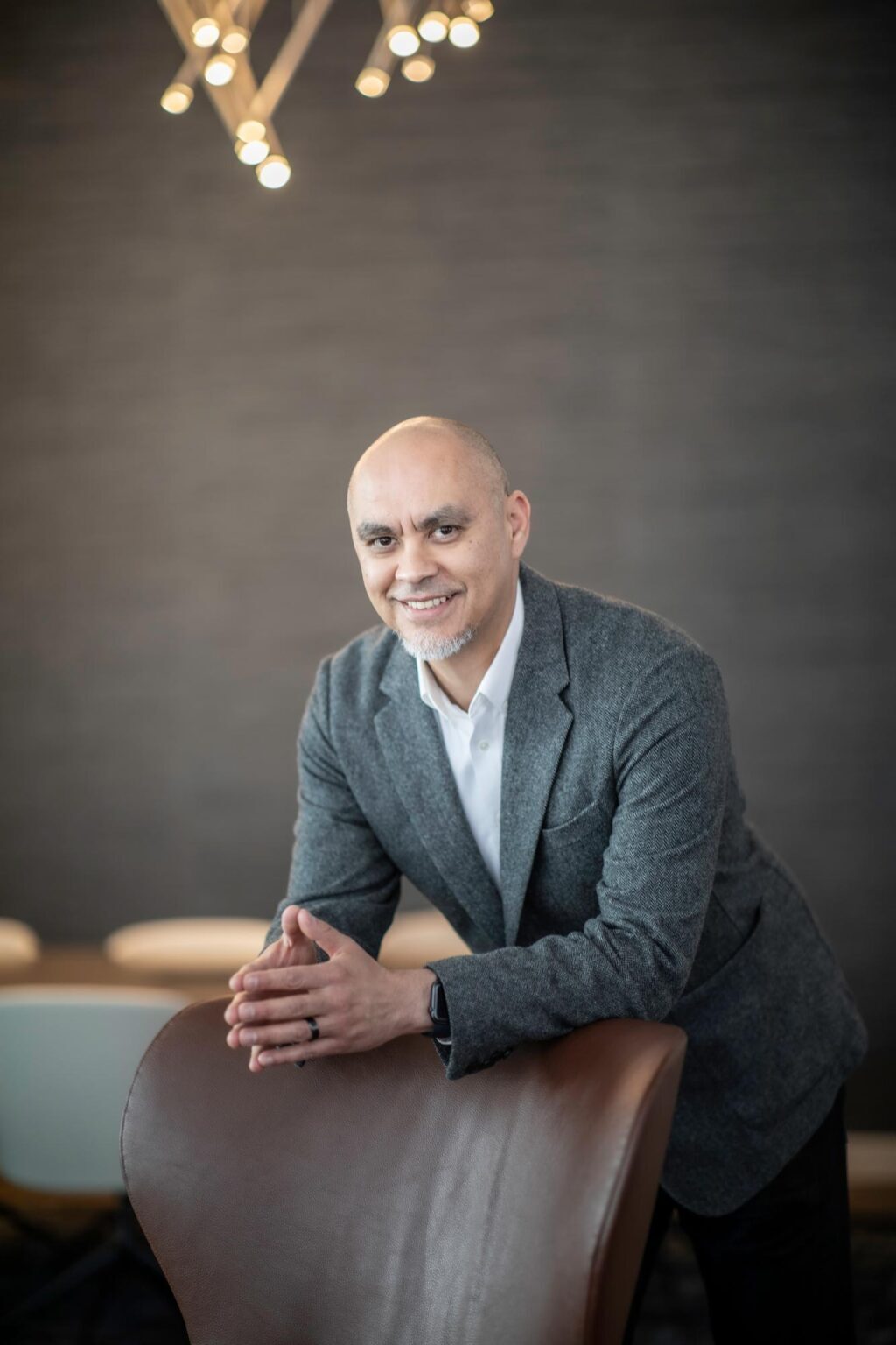 Ricardo Montoya, Principal at GFF, leaning on a brown leather chair in a gray blazer and white shirt beneath modern pendant lighting.