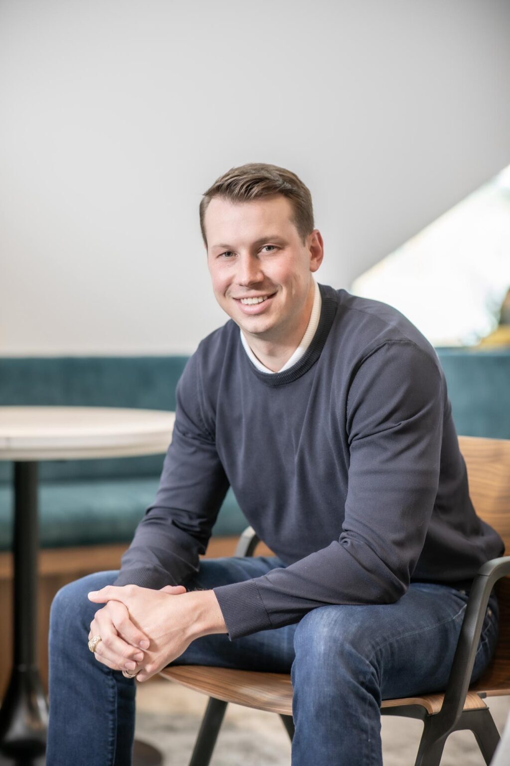 A portrait of Garrett Barker, Associate and Studio Director at GFF, seated in a modern office wearing a dark sweater and jeans, smiling at the camera.