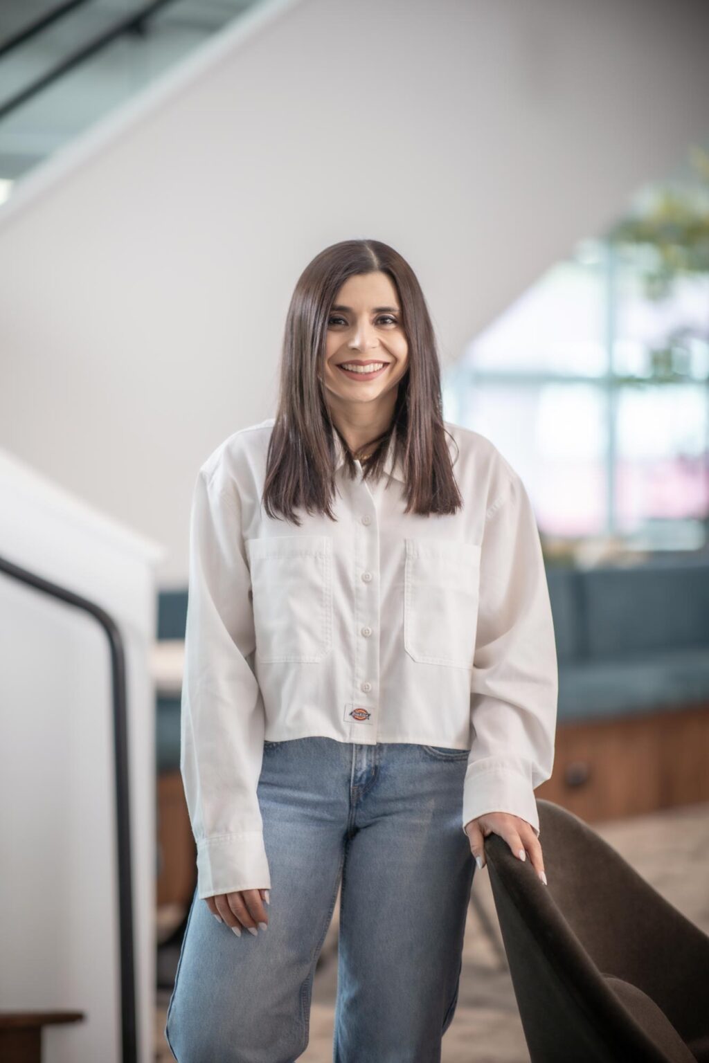 Mariah Trevizo standing in a modern interior space, wearing a white button-up shirt and jeans, smiling near a chair.