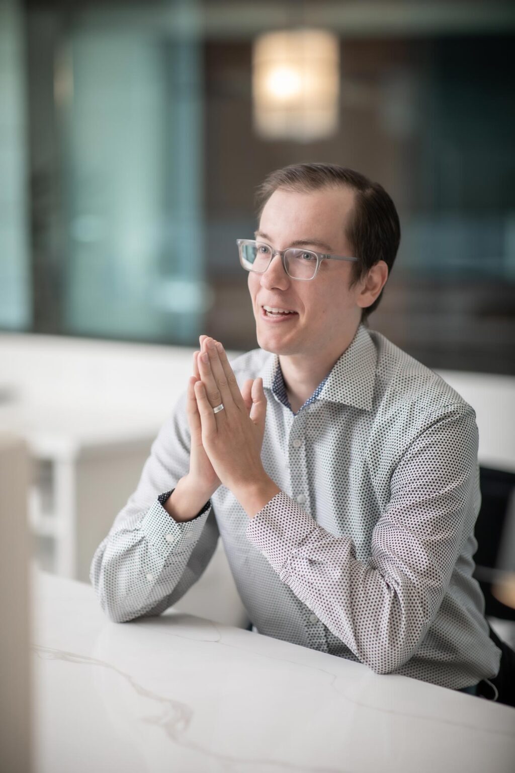 A portrait of Matt Frame, Project Leader at GFF, sitting at a white table in the Dallas office, speaking with hands together while smiling and wearing a patterned shirt and glasses.