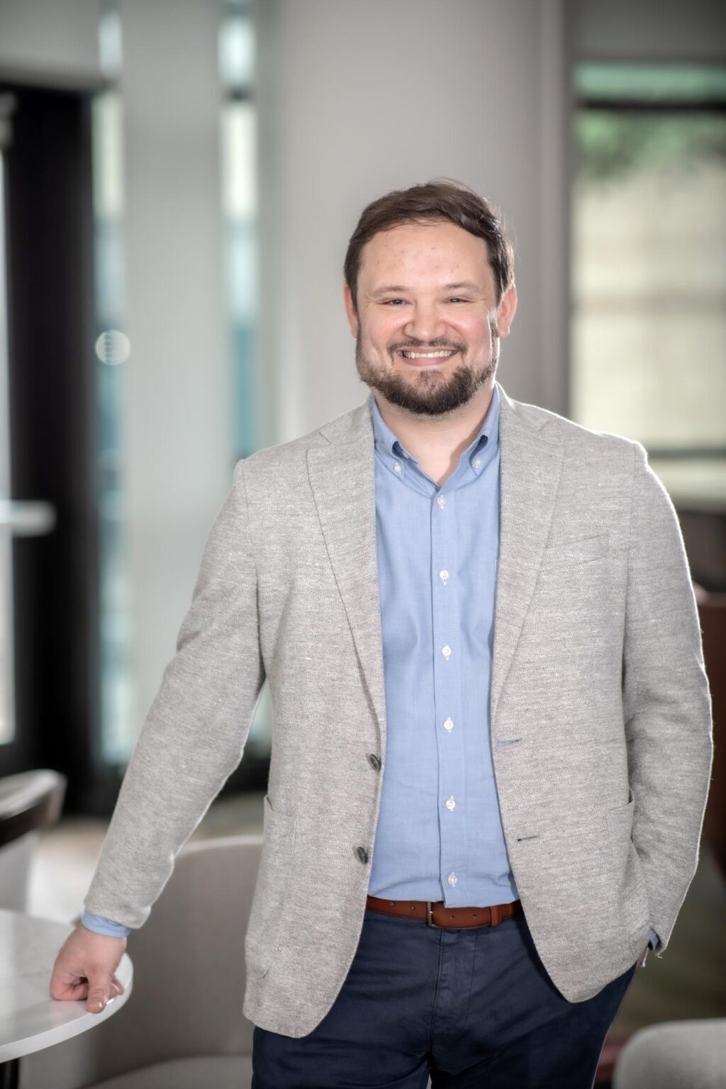 A portrait of Jacob Thibodeaux, Senior Project Coordinator at GFF, standing in an office space wearing a light gray jacket, blue shirt, and navy pants, smiling and resting one hand on a table.