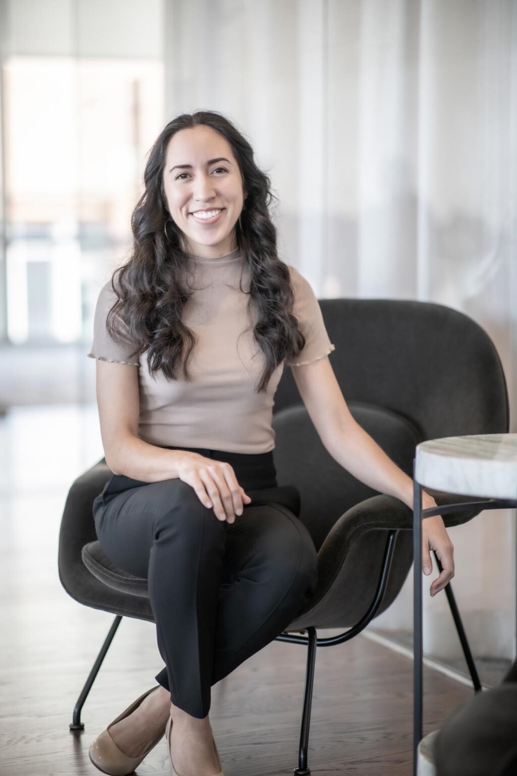 A portrait of Marisa Dominguez, Design Professional at GFF, seated in a modern office lounge chair, smiling and wearing a beige top with black pants.