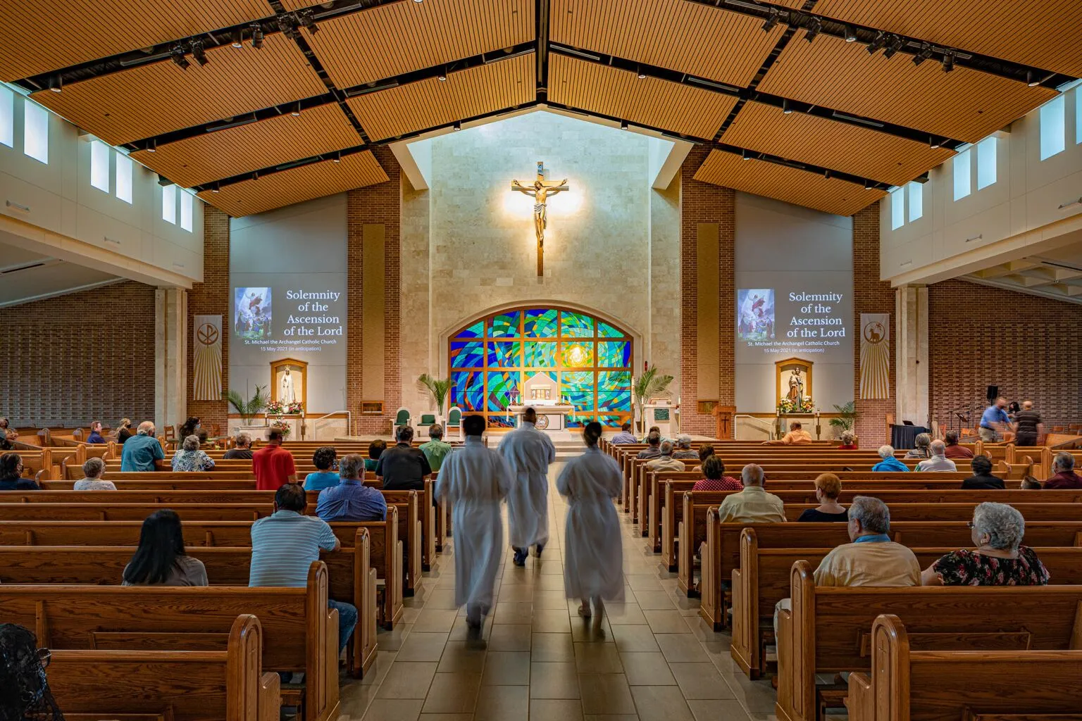 Rear view of the nave showcasing fan-shaped seating, stained glass at the altar, and a warm wood ceiling.