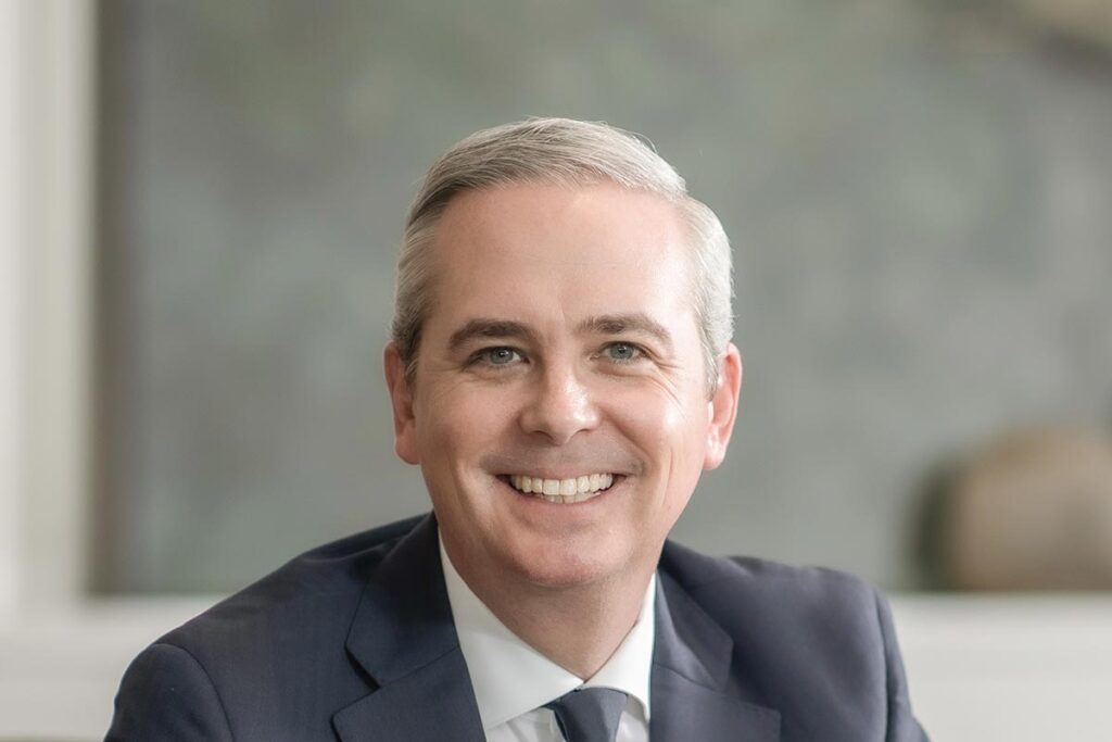Close-up portrait of Evan Beattie smiling warmly in a professional office environment, wearing a navy suit and tie.