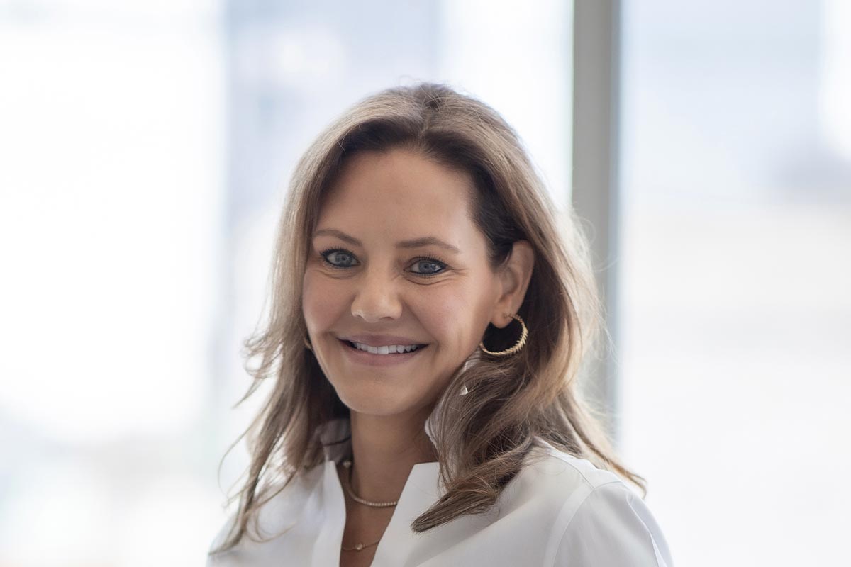 Close-up portrait of Allison Hubbard in front of a softly blurred background, wearing a white blouse and gold hoop earrings.