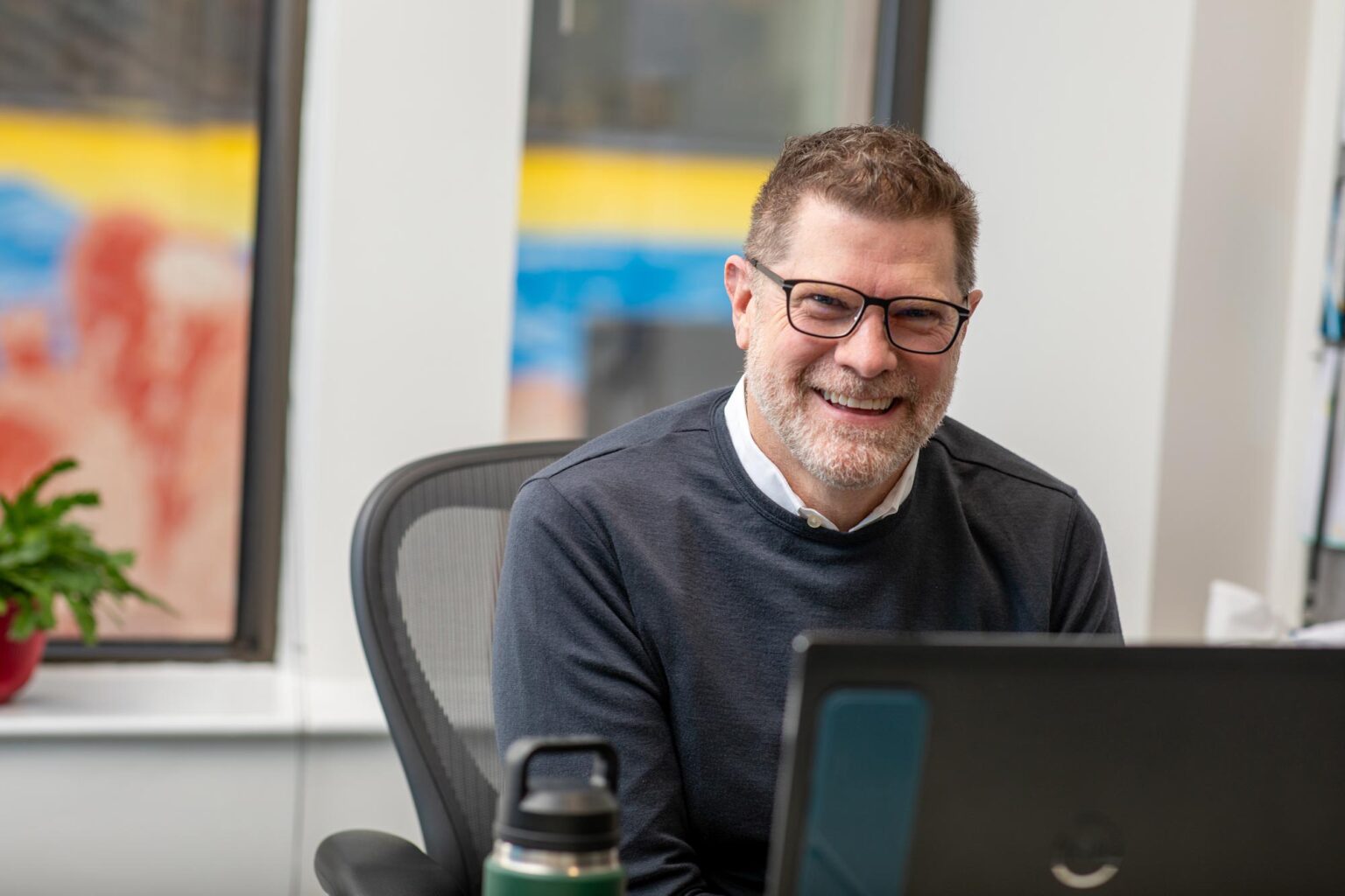 Jim West, AIA, LEED AP, NCARB, Principal and Managing Director at GFF Austin, seated at a desk with a laptop, smiling in a bright modern office with colorful mural outside the window.
