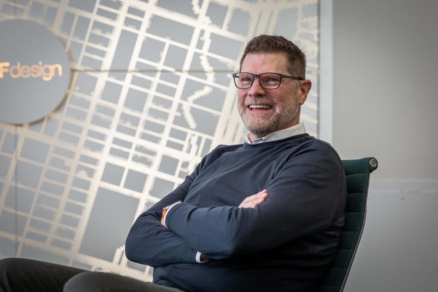 Jim West, AIA, LEED AP, NCARB, Principal and Managing Director at GFF Austin, seated with arms crossed in front of a wall graphic, smiling during a candid office moment.
