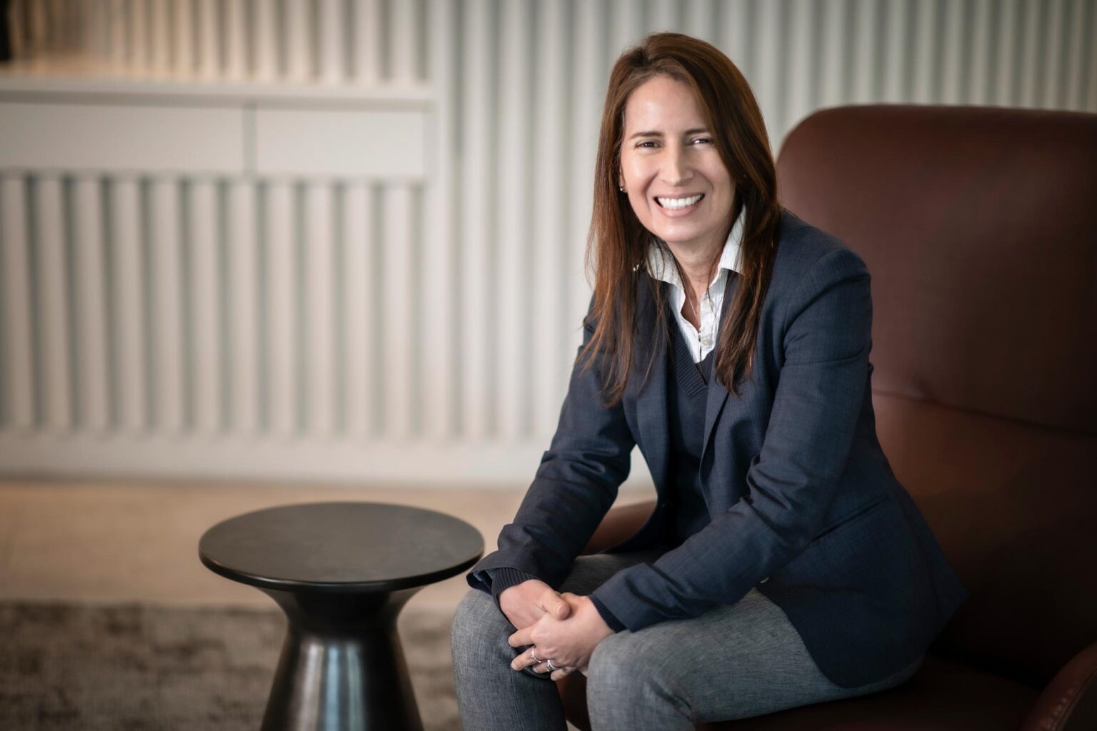 Maria Gomez, AIA, LEED AP, Principal and Studio Director at GFF, seated in a leather chair wearing a navy blazer and gray pants, smiling in a modern office setting.