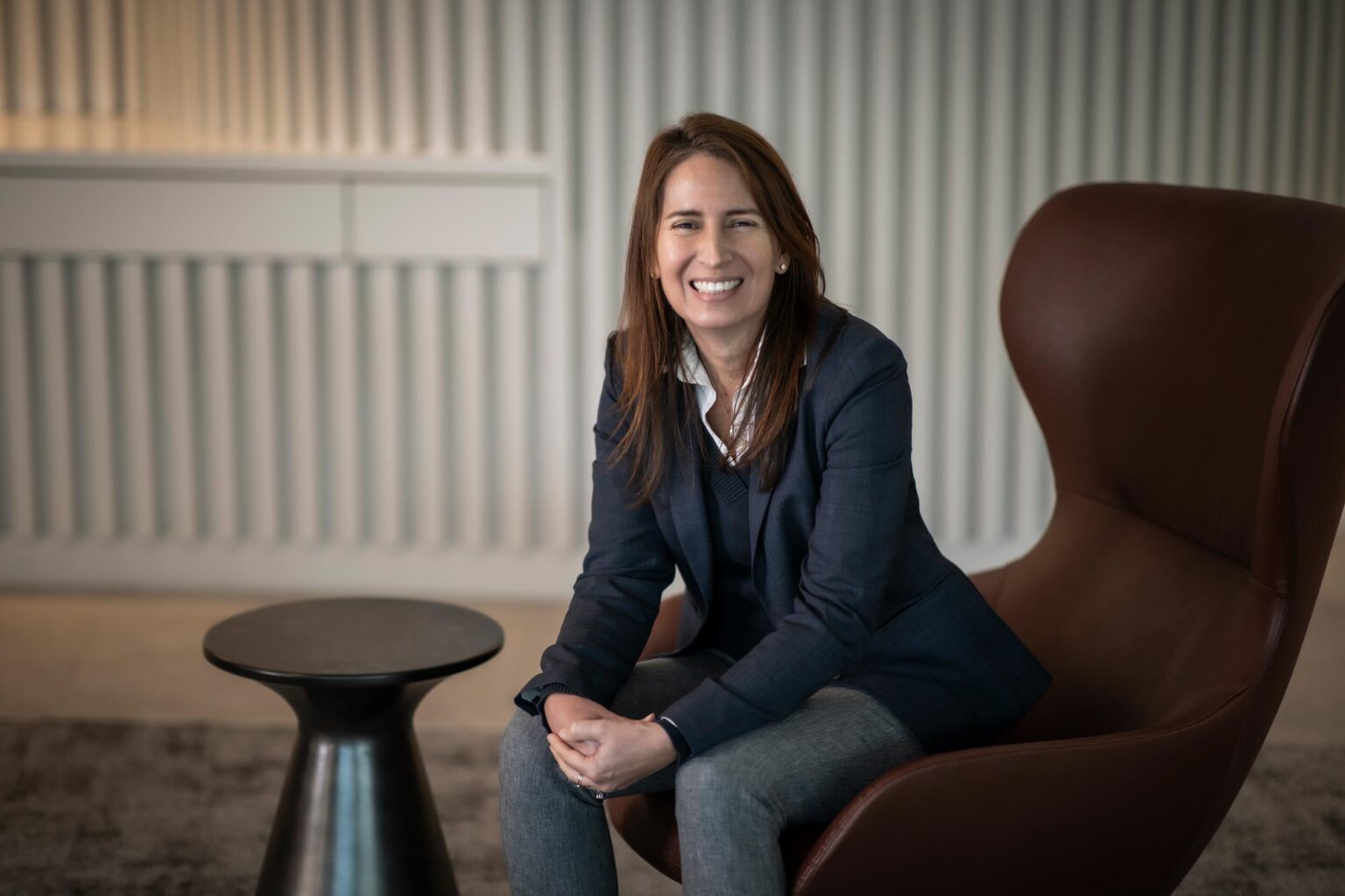 Maria Gomez, AIA, LEED AP, Principal and Studio Director at GFF, seated in a modern office lounge chair with folded hands, smiling confidently.