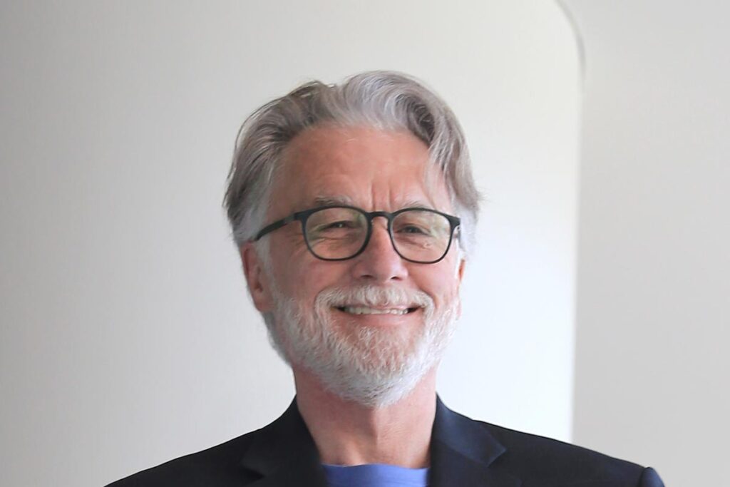 Close-up portrait of Brian Kuper wearing glasses and a dark jacket, smiling in front of a light background.