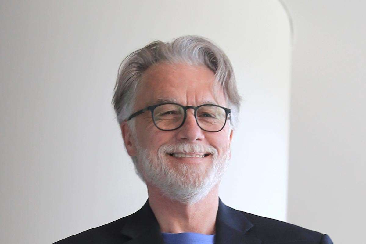 Close-up portrait of Brian Kuper wearing glasses and a dark jacket, smiling in front of a light background.