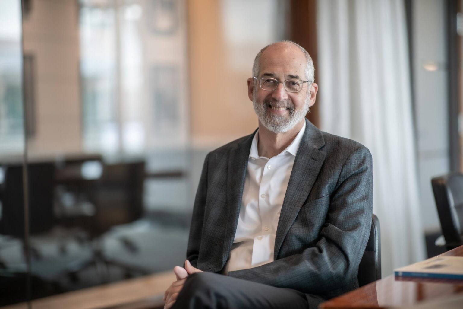 Jon Rollins, Principal at GFF, seated in a conference room with glass walls, wearing a checkered gray jacket and white shirt, smiling at camera.