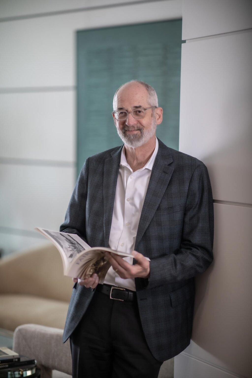 Jon Rollins, Principal at GFF, leaning against wall with open architecture book, dressed in gray jacket and white shirt, photographed in a light-filled office.
