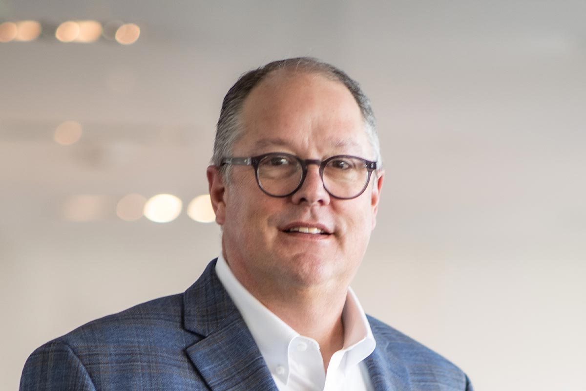 Close-up portrait of John Shiver smiling, wearing glasses and a blue plaid blazer in front of a softly blurred background.