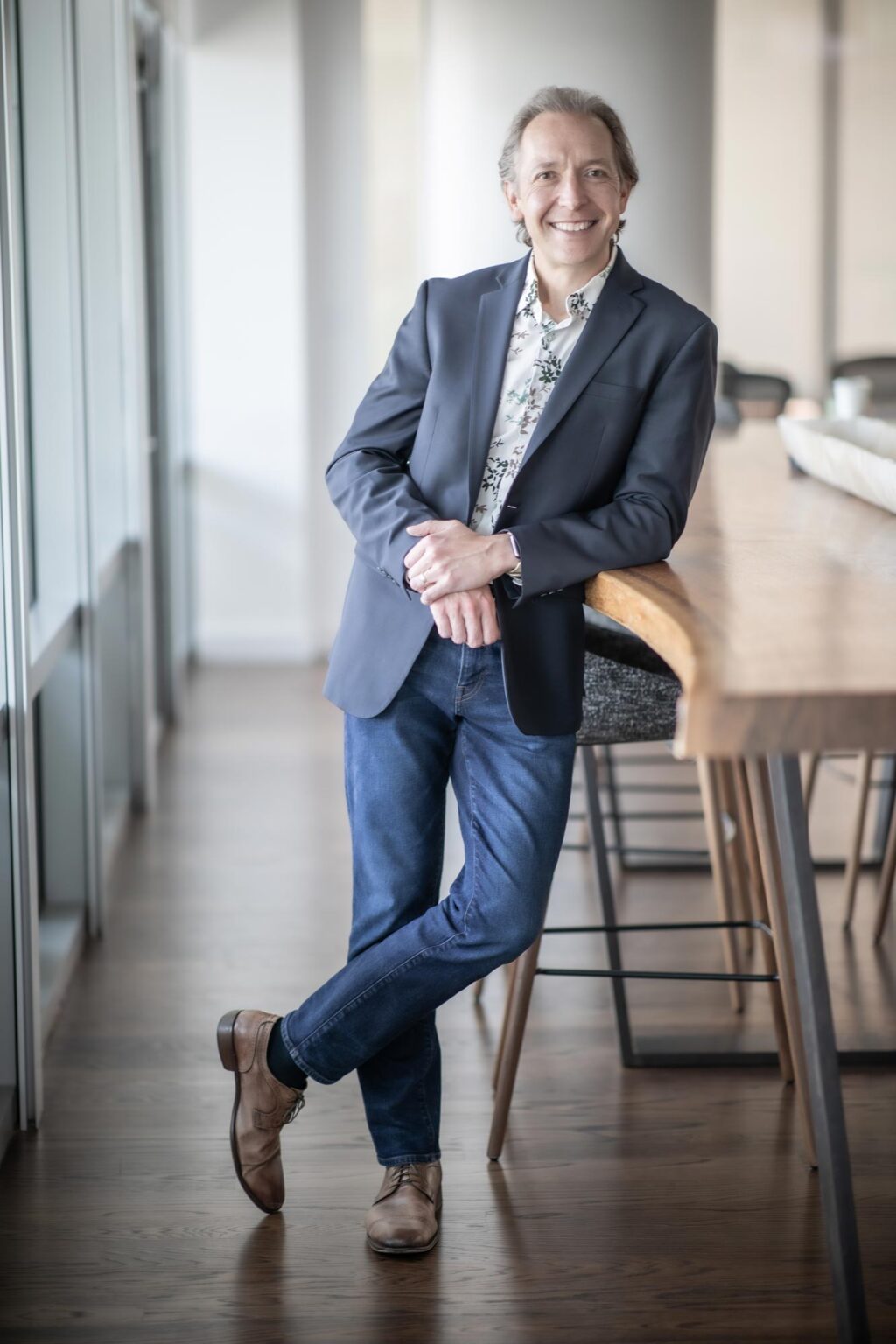 A portrait of Michael Lehr, Associate Principal and Studio Director at GFF, standing in a bright office corridor leaning against a wood counter, wearing a navy jacket and patterned shirt.