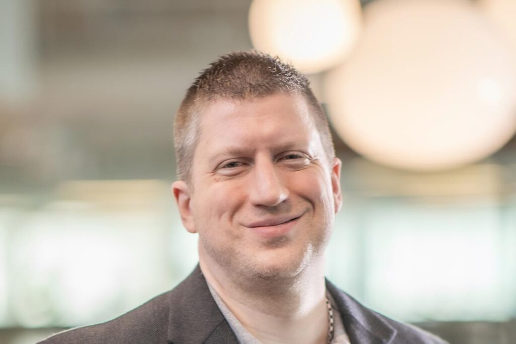 Close-up portrait of Phil DeLaughter wearing a gray blazer, smiling in front of a softly lit office background.