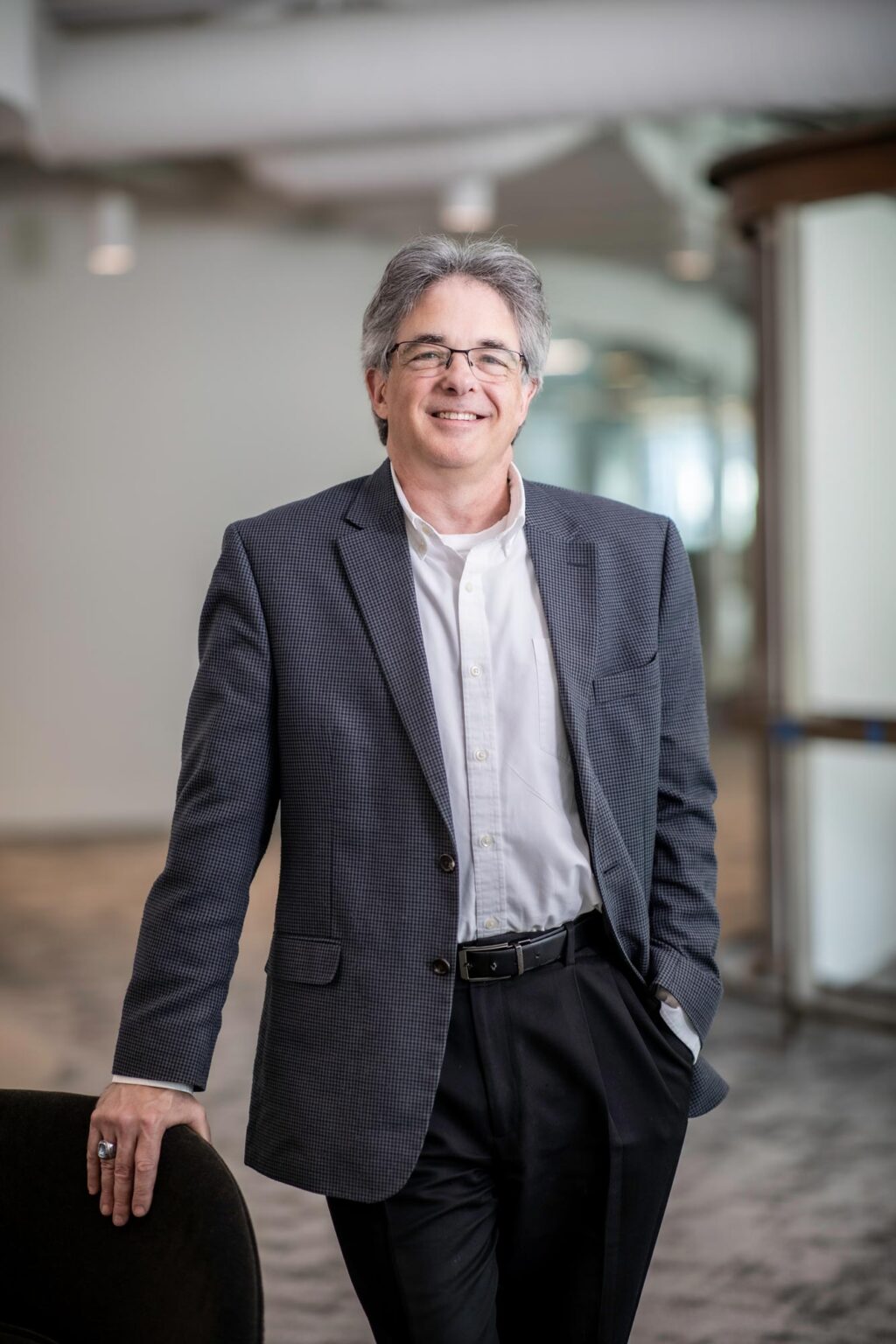 A portrait of Scott Kanaga, Associate Principal and Studio Director at GFF, standing in the Dallas office wearing a dark patterned jacket and white shirt, smiling with one hand resting on a chair.