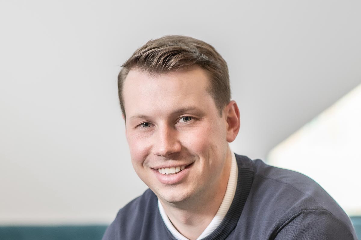 A cropped landscape portrait of Garrett Barker, Associate and Studio Director at GFF, smiling in a softly lit office environment.