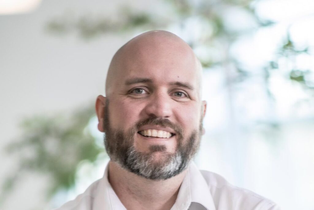 Close-up portrait of Jason Hoyt wearing a white shirt and smiling, with a softly blurred office background.