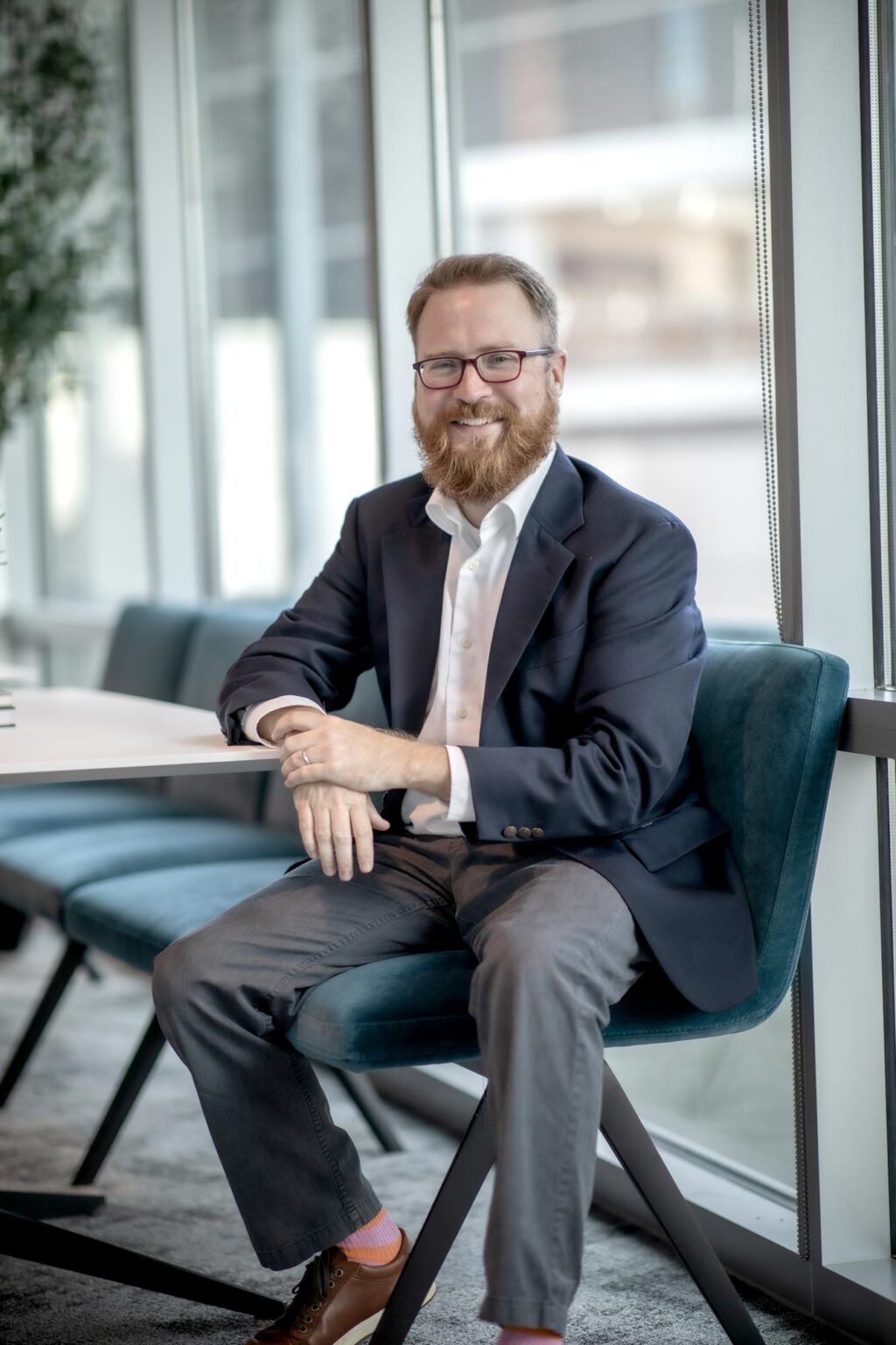 A portrait of Sam Stribling, Associate and Studio Director at GFF, seated in a modern office with large windows and teal seating.