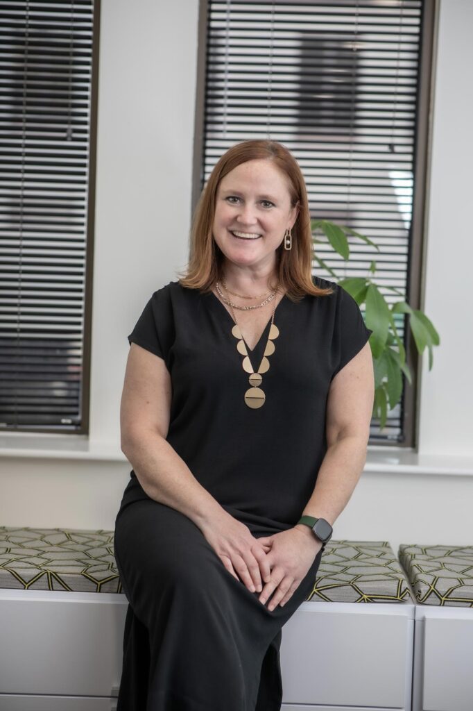 Sara Good, Associate and Studio Director at GFF, sitting near a window in a black dress with geometric-patterned bench and indoor plant in the background.