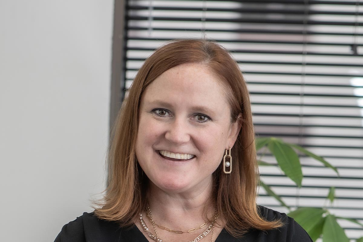 Sara Good, Associate and Studio Director at GFF, close-up portrait with window blinds and plant in background, wearing gold jewelry and black blouse.