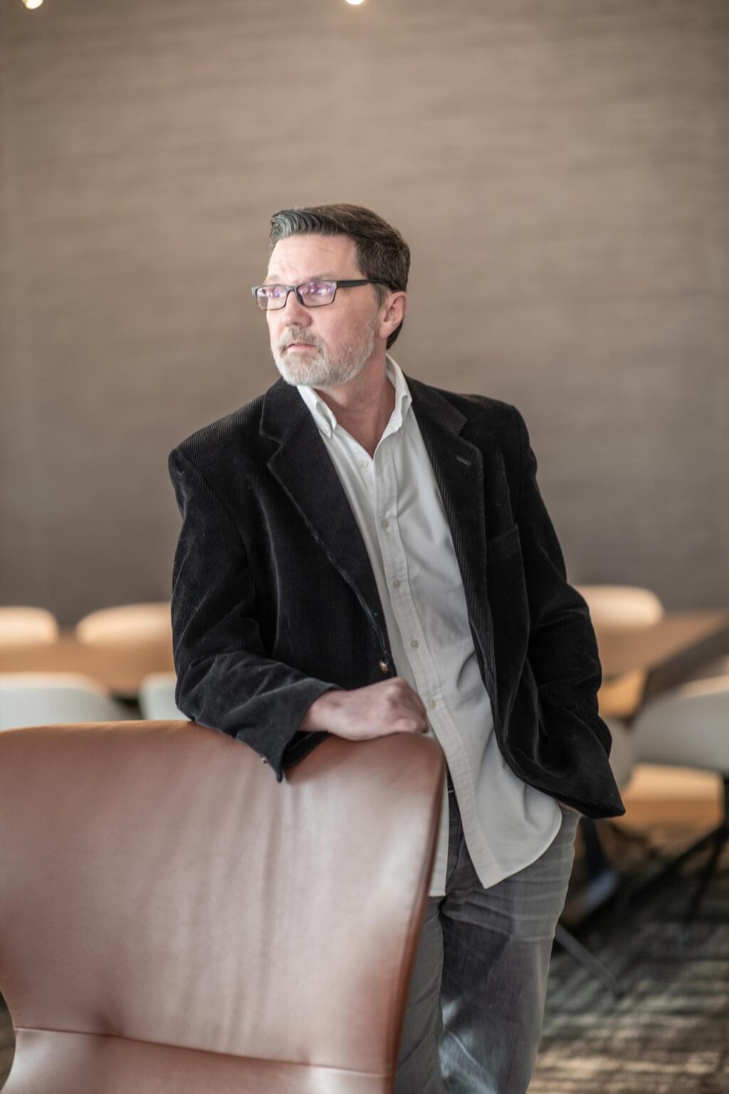 Todd Toupal standing in a modern meeting space wearing a black jacket and white shirt, looking thoughtfully to the side.