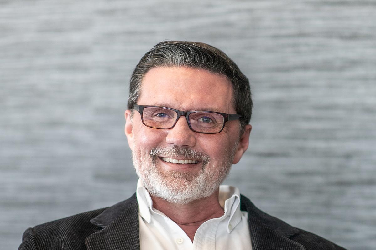 Close-up portrait of Todd Toupal smiling warmly in front of a textured gray background.