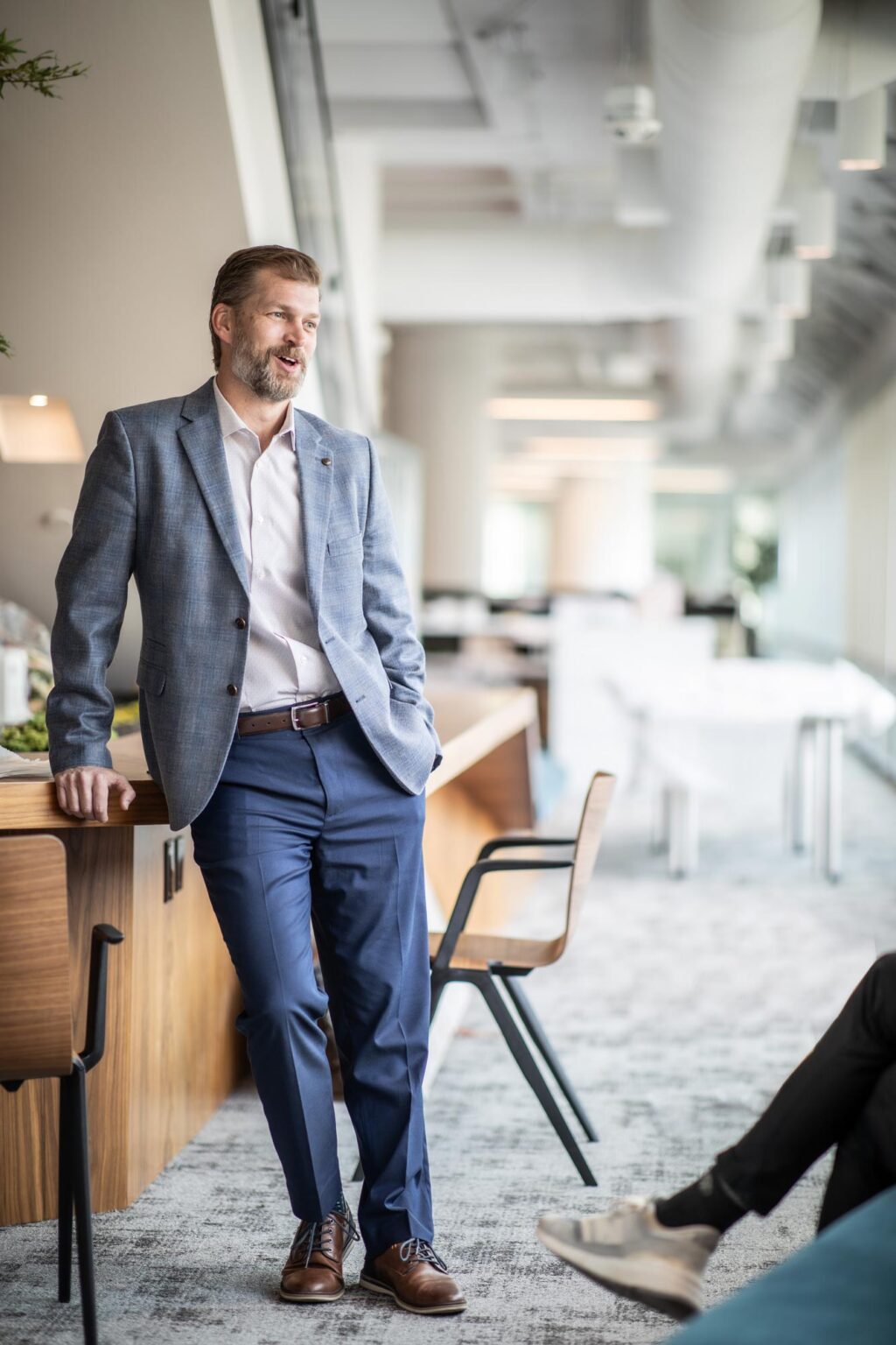 Grant Wickard, AIA, Associate Principal and Senior Project Leader at GFF, standing and speaking with colleagues in a modern office lounge while wearing a gray blazer and blue slacks.