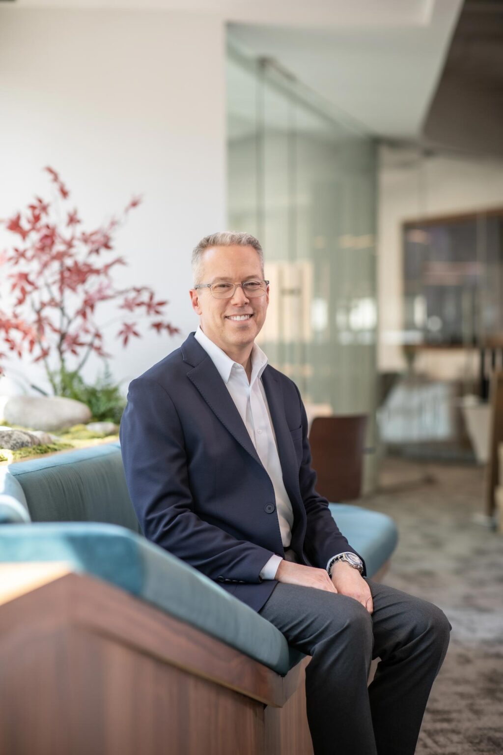 Russell Hagg seated in a bright office interior with natural light and plants in the background, wearing a navy jacket and white shirt.