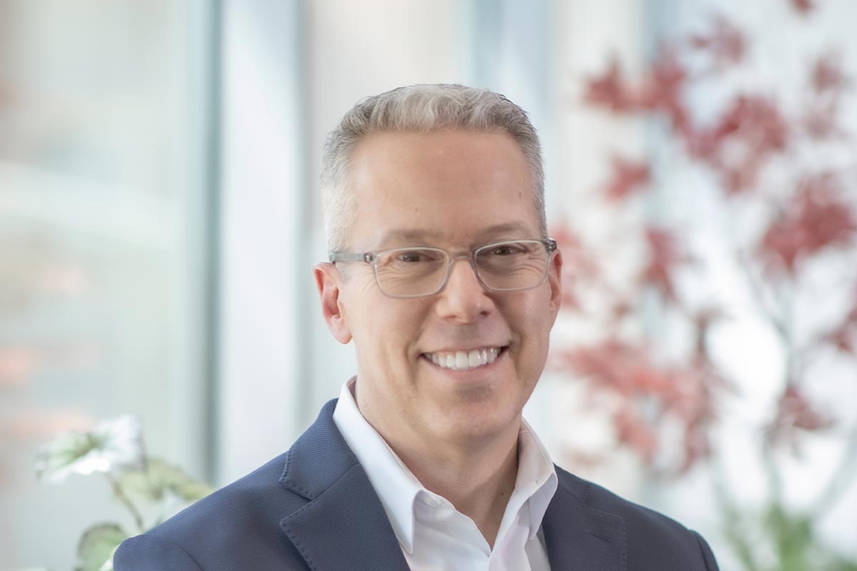 Close-up portrait of Russell Hagg wearing glasses and a navy jacket, smiling in front of a softly blurred office background with plants.