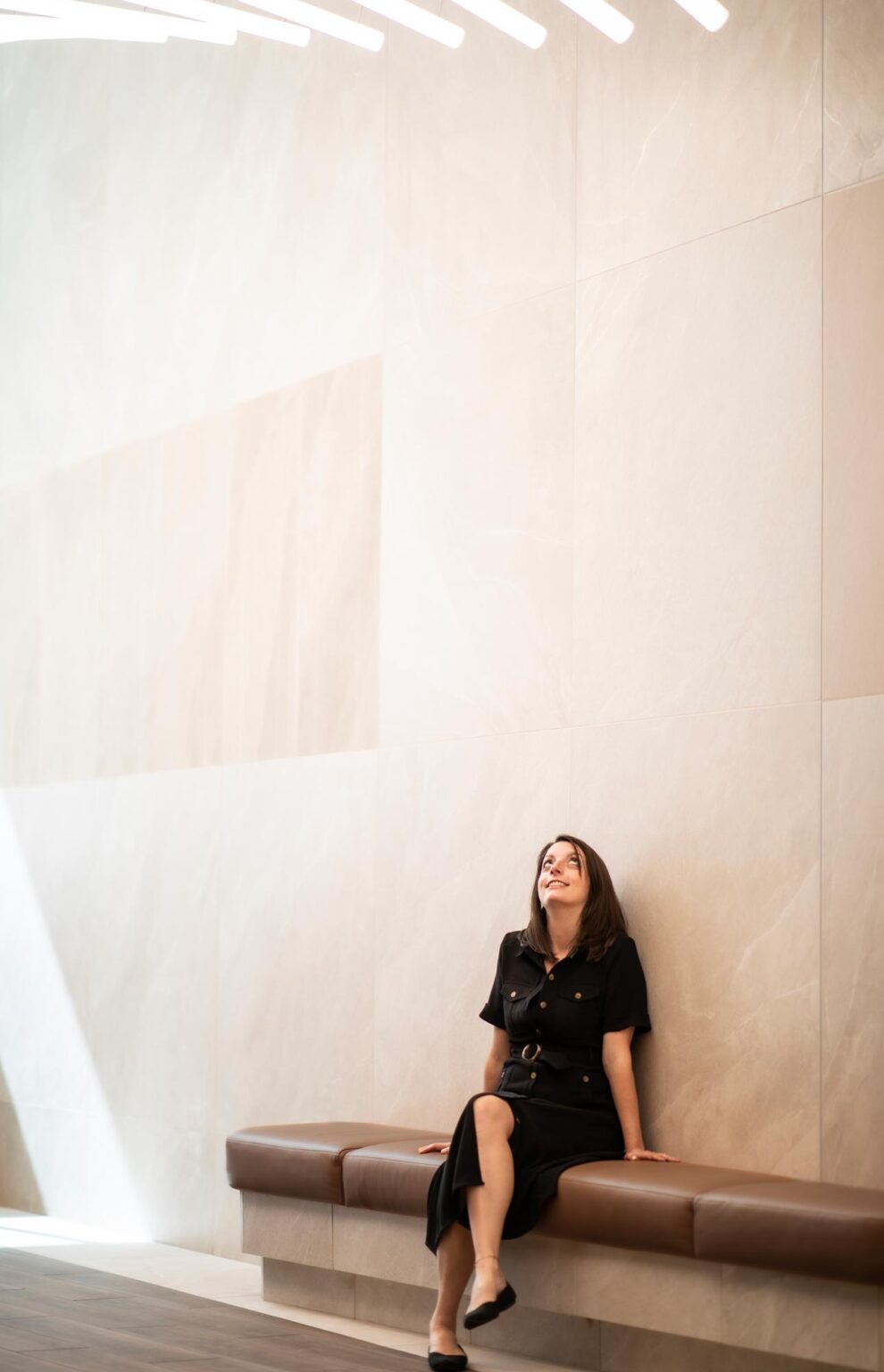 Beth Schmidt, RID, Associate and Senior Design Leader at GFF, seated on a bench in a tall stone-clad lobby, looking upward toward architectural lighting above.