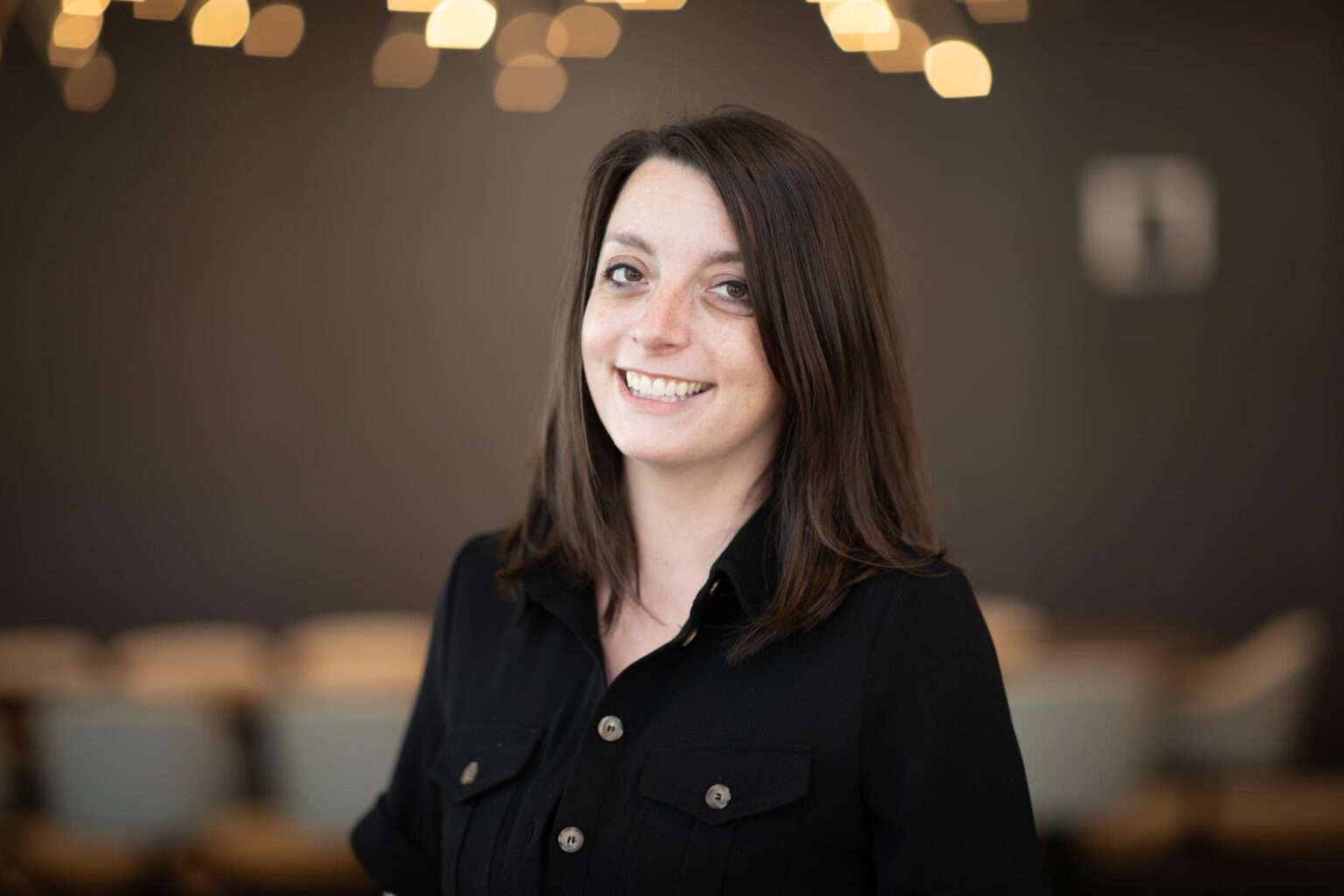 Beth Schmidt, RID, Associate and Senior Design Leader at GFF, smiling in a black dress with warm background lights softly blurred behind her.
