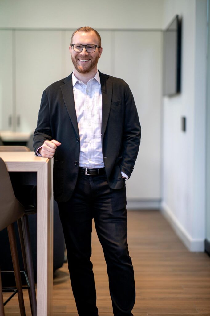 Blake Thames, Associate and Senior Design Leader at GFF, standing beside a tall table in a tailored black jacket and checkered shirt, smiling confidently.