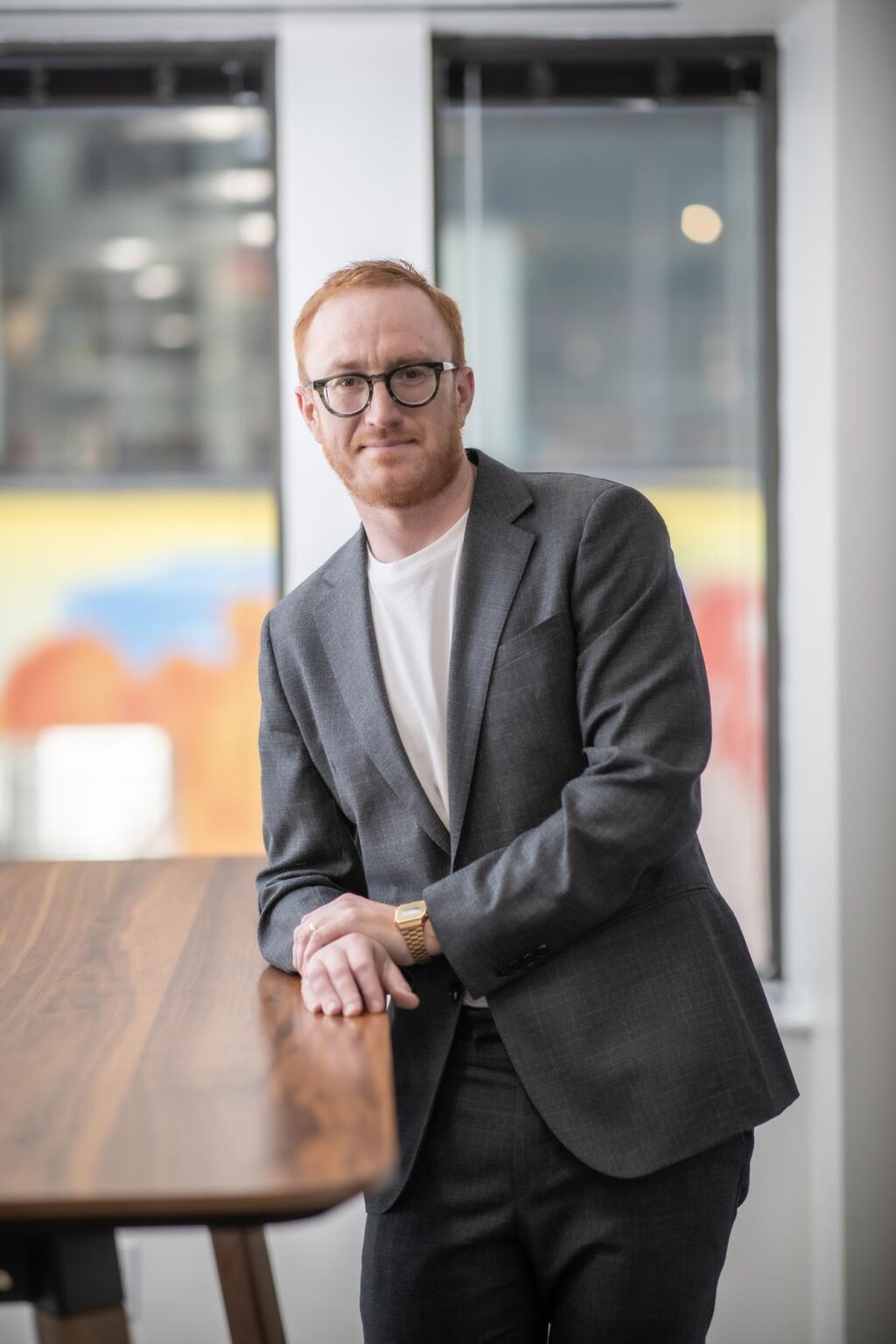 Christopher Day, Associate and Senior Design Leader at GFF, leaning on a wooden table in a gray suit and white shirt in a modern office.