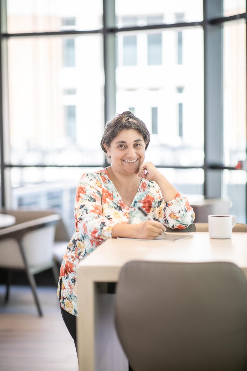 A portrait of Nardine Mansi, Associate and Senior Project Leader at GFF, seated at a high table with a notebook and coffee mug in a sunlit workspace.