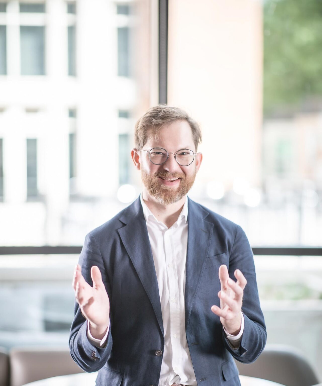 Gavin Newman seated in a bright office, gesturing while speaking and smiling, dressed in a navy blazer and white shirt.