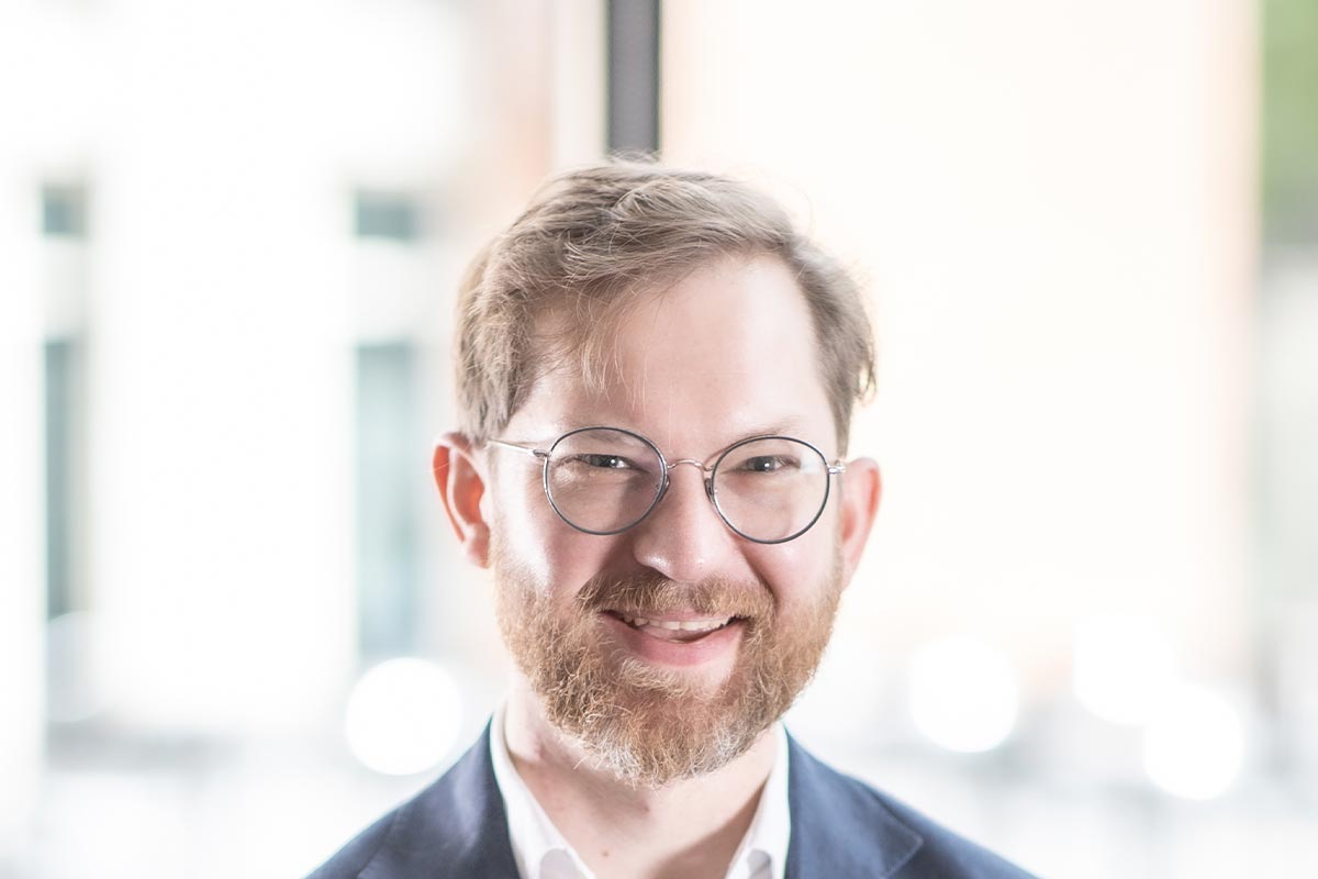 Close-up portrait of Gavin Newman wearing glasses and a navy blazer, smiling in front of a bright background.