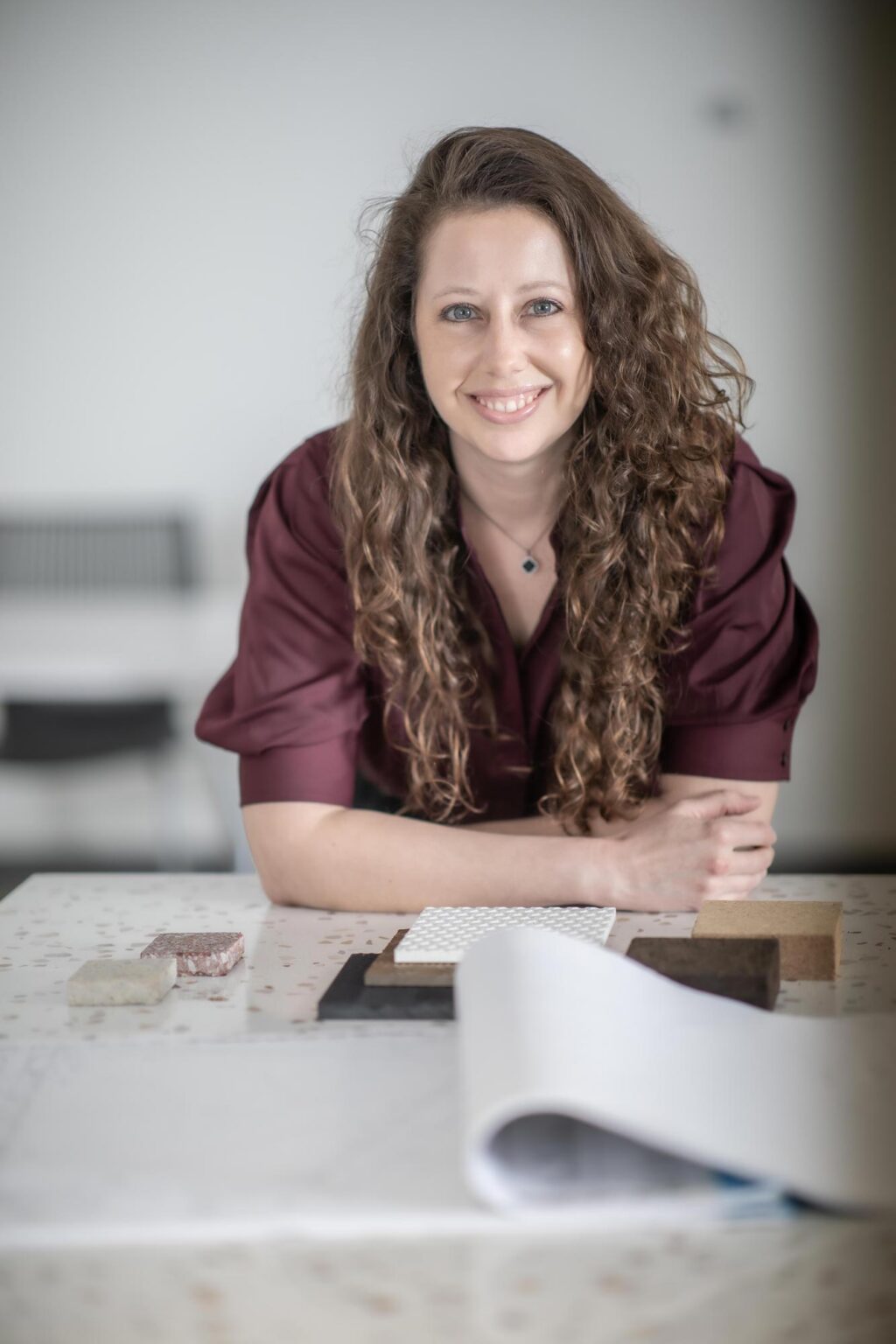 Jamie Wallace leaning on a materials table with samples and drawings, wearing a maroon blouse and smiling in a bright studio setting.