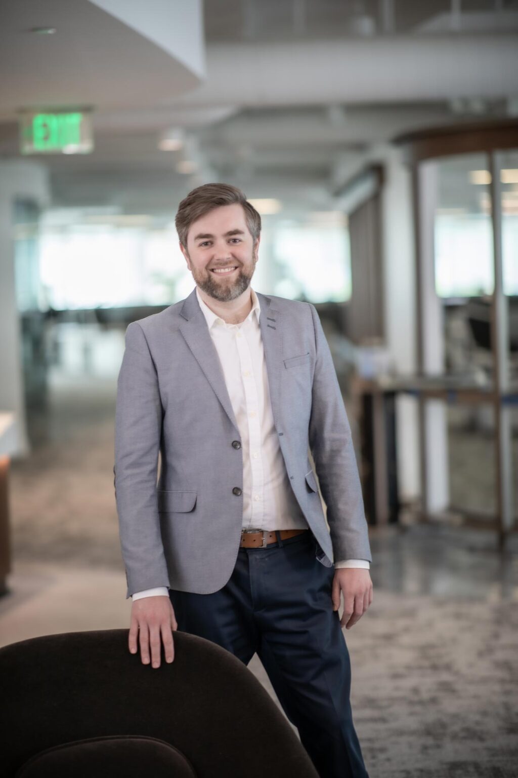 A portrait of Josh Frederick, Associate and Project Leader at GFF, standing in the Dallas office wearing a light gray jacket and white shirt, smiling with one hand resting on a chair.