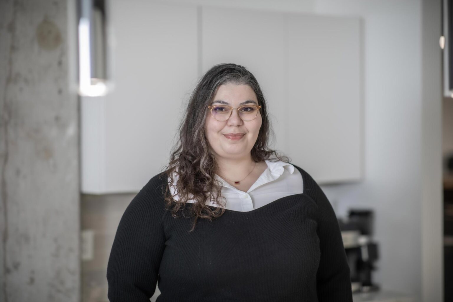 Sarah Hamzeh standing in a contemporary office setting, wearing a black sweater over a white blouse and smiling warmly.