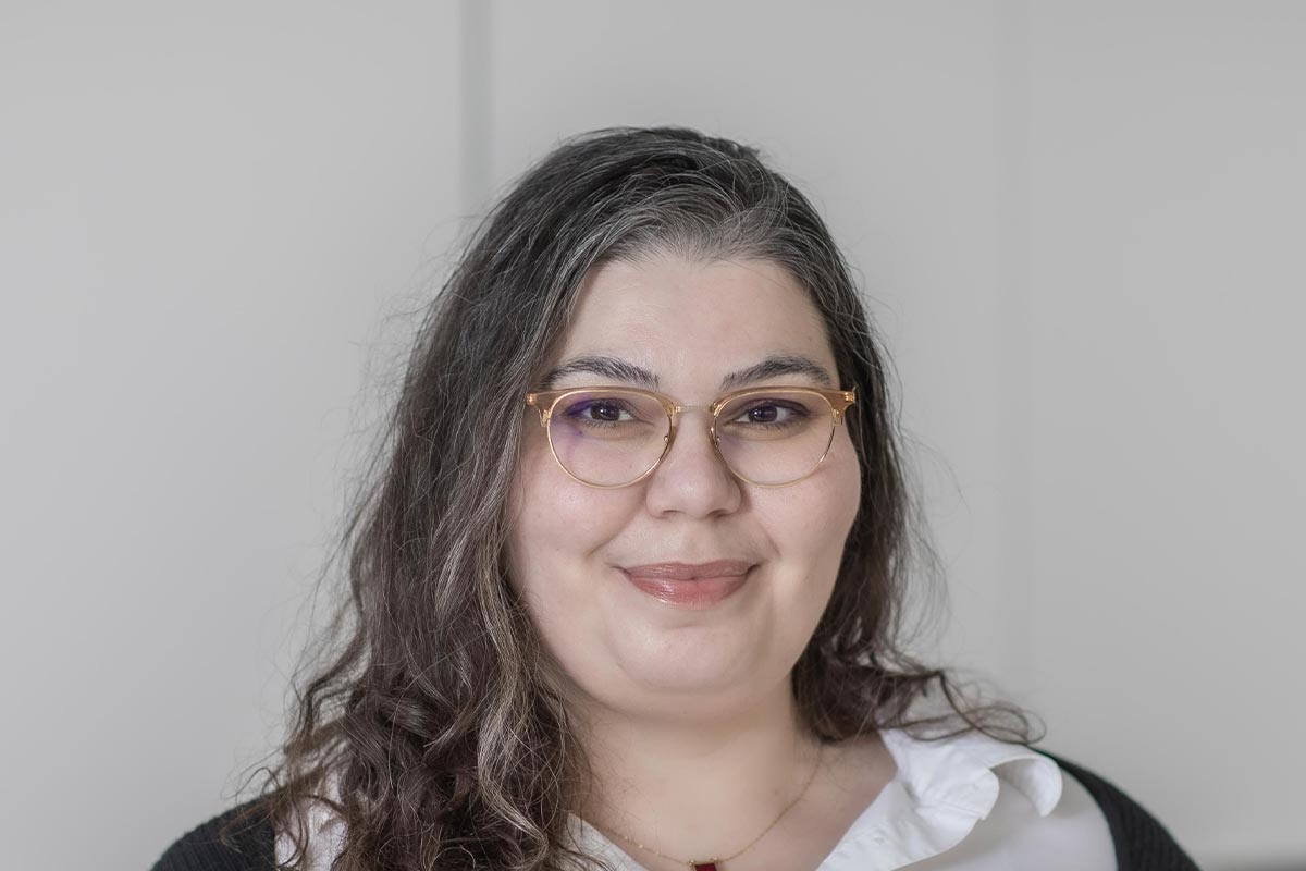 Close-up portrait of Sarah Hamzeh wearing glasses and a white blouse, smiling in front of a softly lit neutral background.