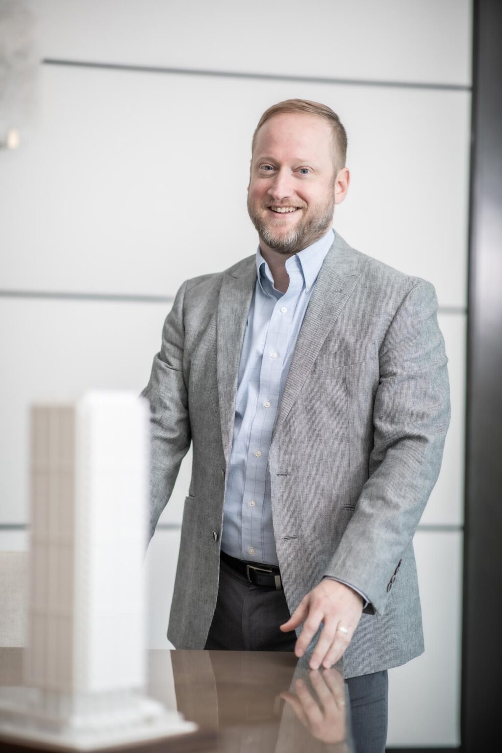 A portrait of David Anderson, Senior Project Leader at GFF, standing beside an architectural model in a bright office with a white wall background, wearing a light gray jacket and blue shirt.