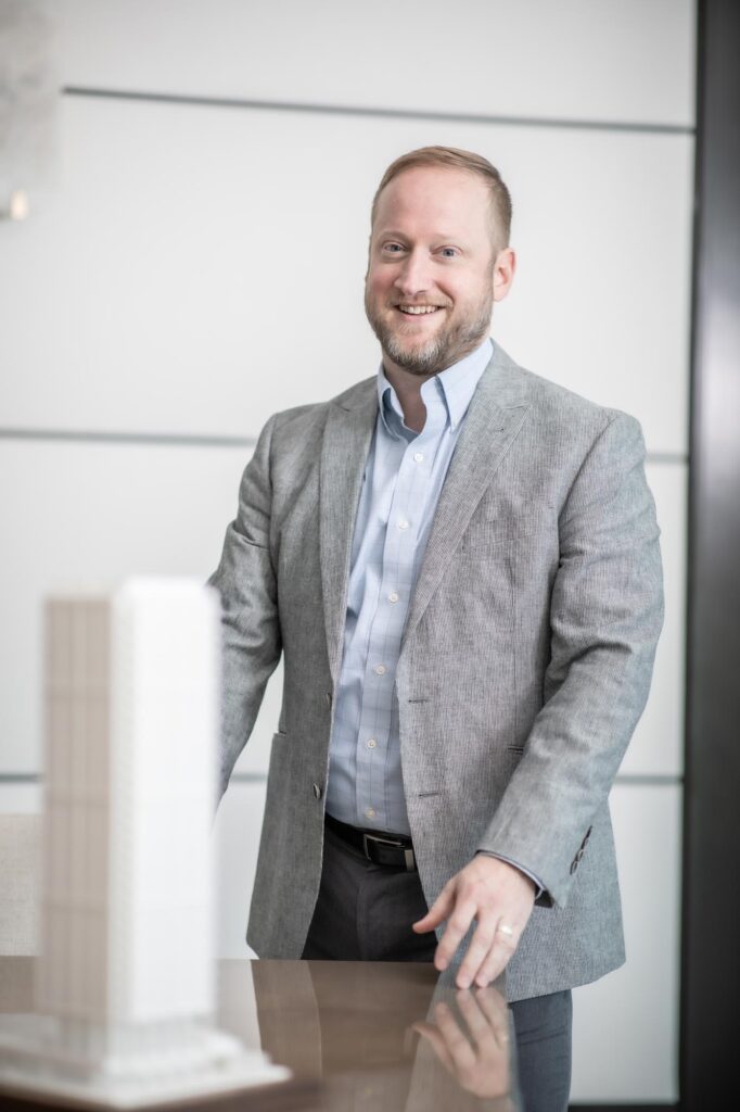 A portrait of David Anderson, Senior Project Leader at GFF, standing beside an architectural model in a bright office with a white wall background, wearing a light gray jacket and blue shirt.
