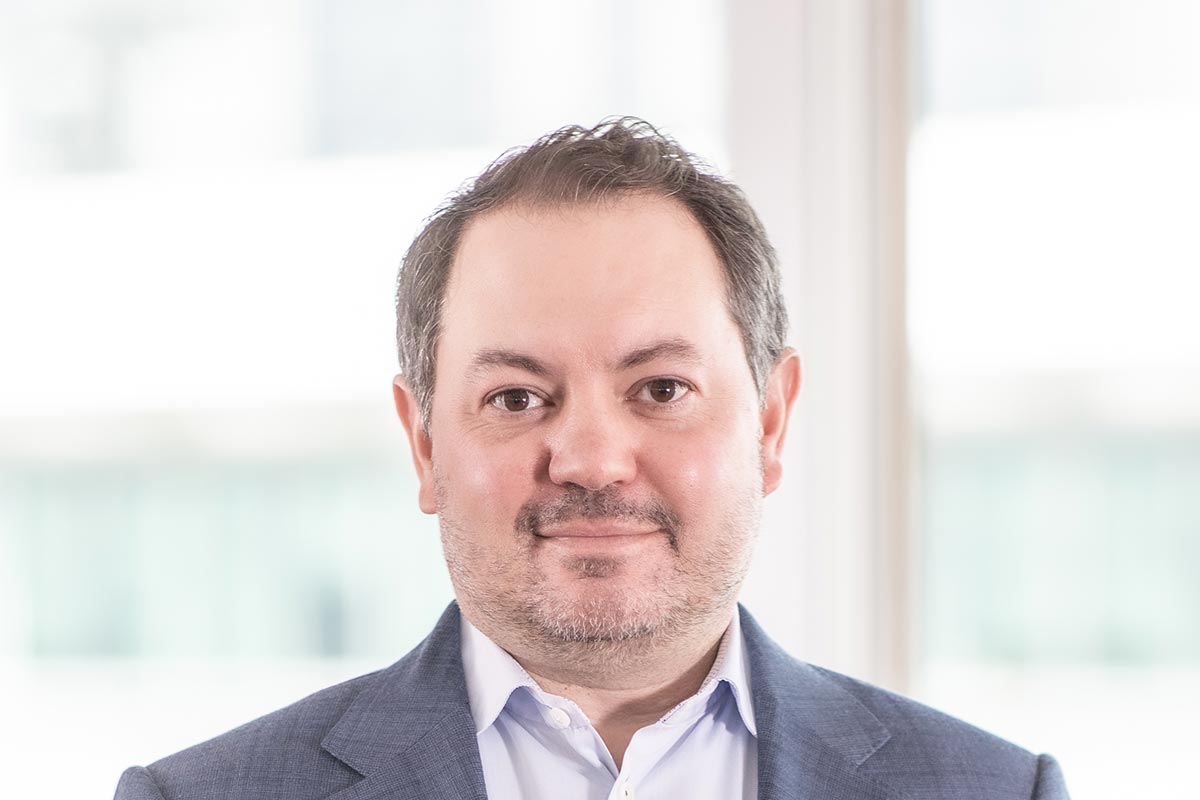 Close-up portrait of David Rosenbloom wearing a blue blazer and white shirt, in front of a bright office window background.
