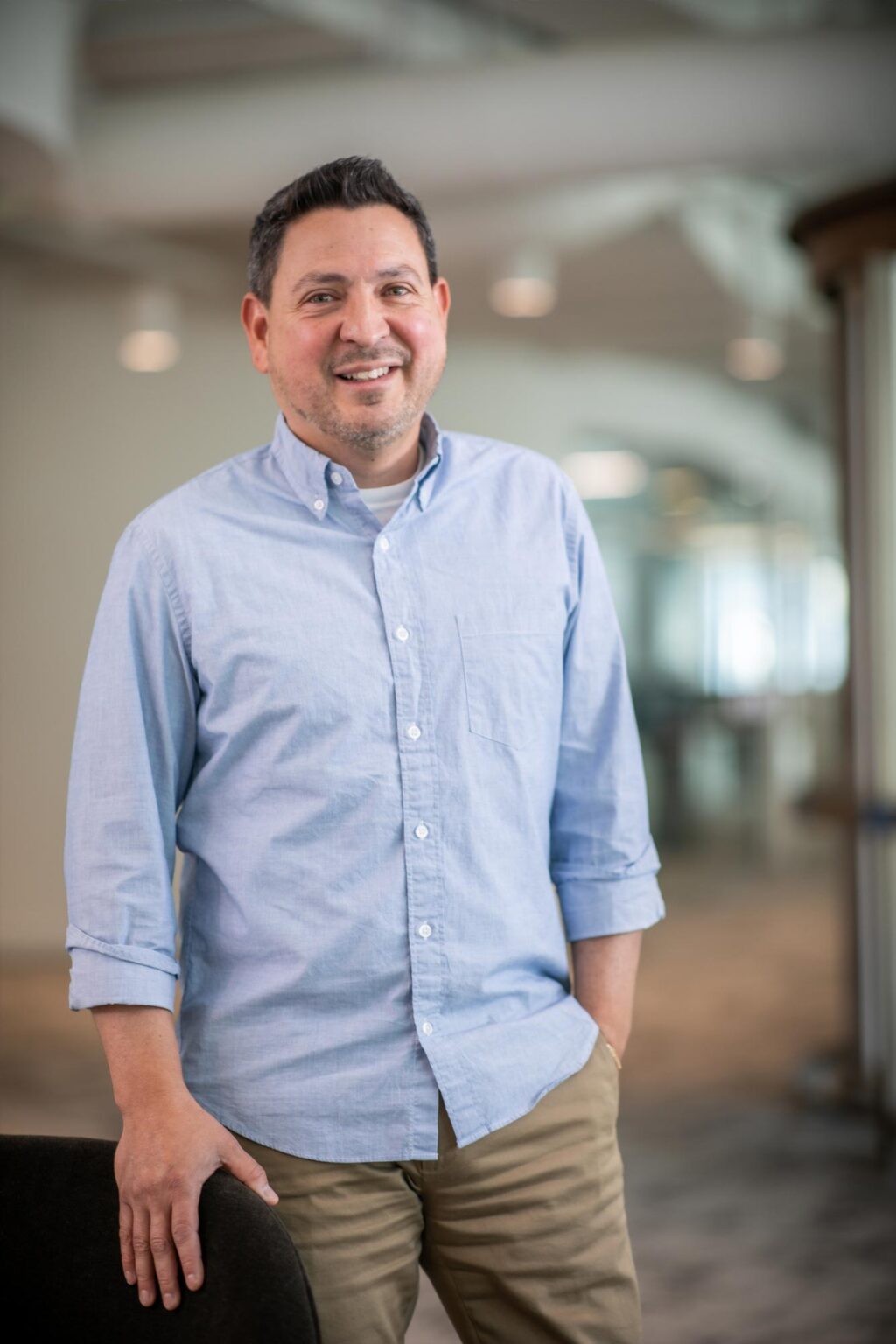 Eric Marquez de la Plata standing and smiling in a bright office corridor, wearing a light blue button-down shirt and khaki pants.