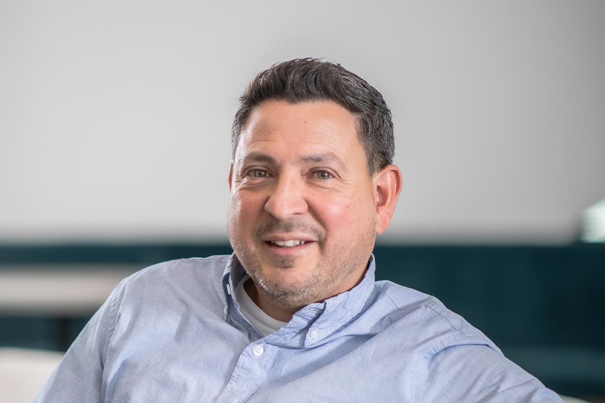Close-up portrait of Eric Marquez de la Plata smiling in a light blue shirt, seated in a softly lit modern workspace.