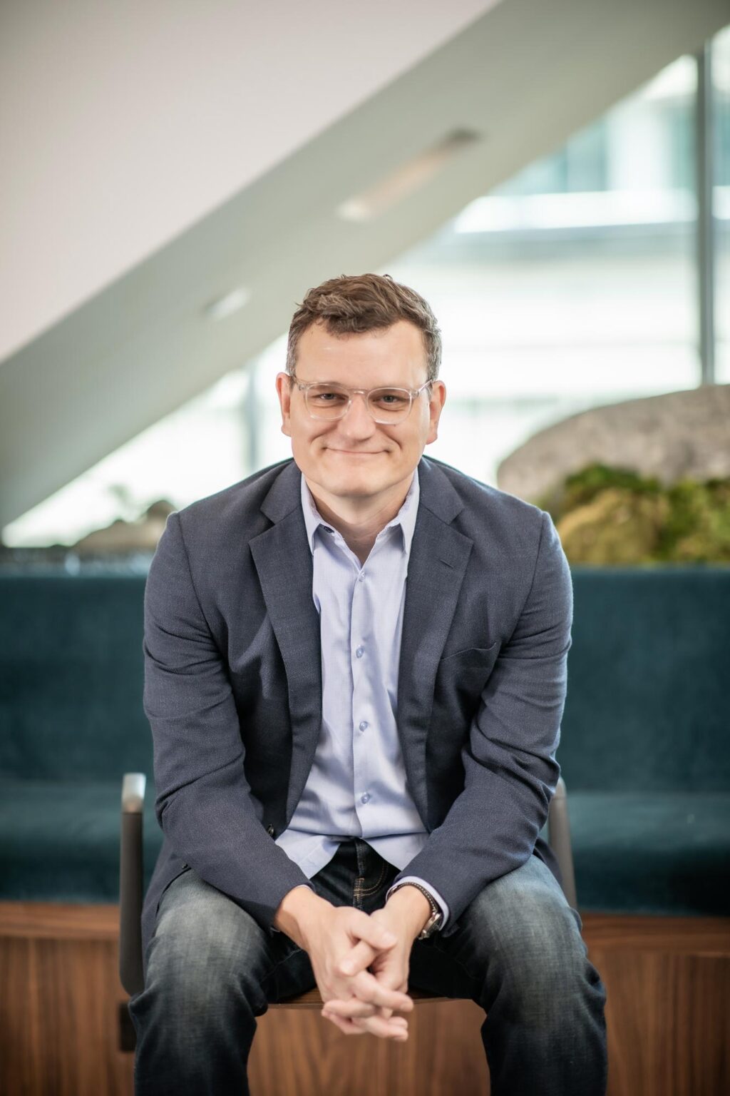 John Jacobson seated in a modern interior with a sloped ceiling and natural light, wearing a dark blazer and light shirt.