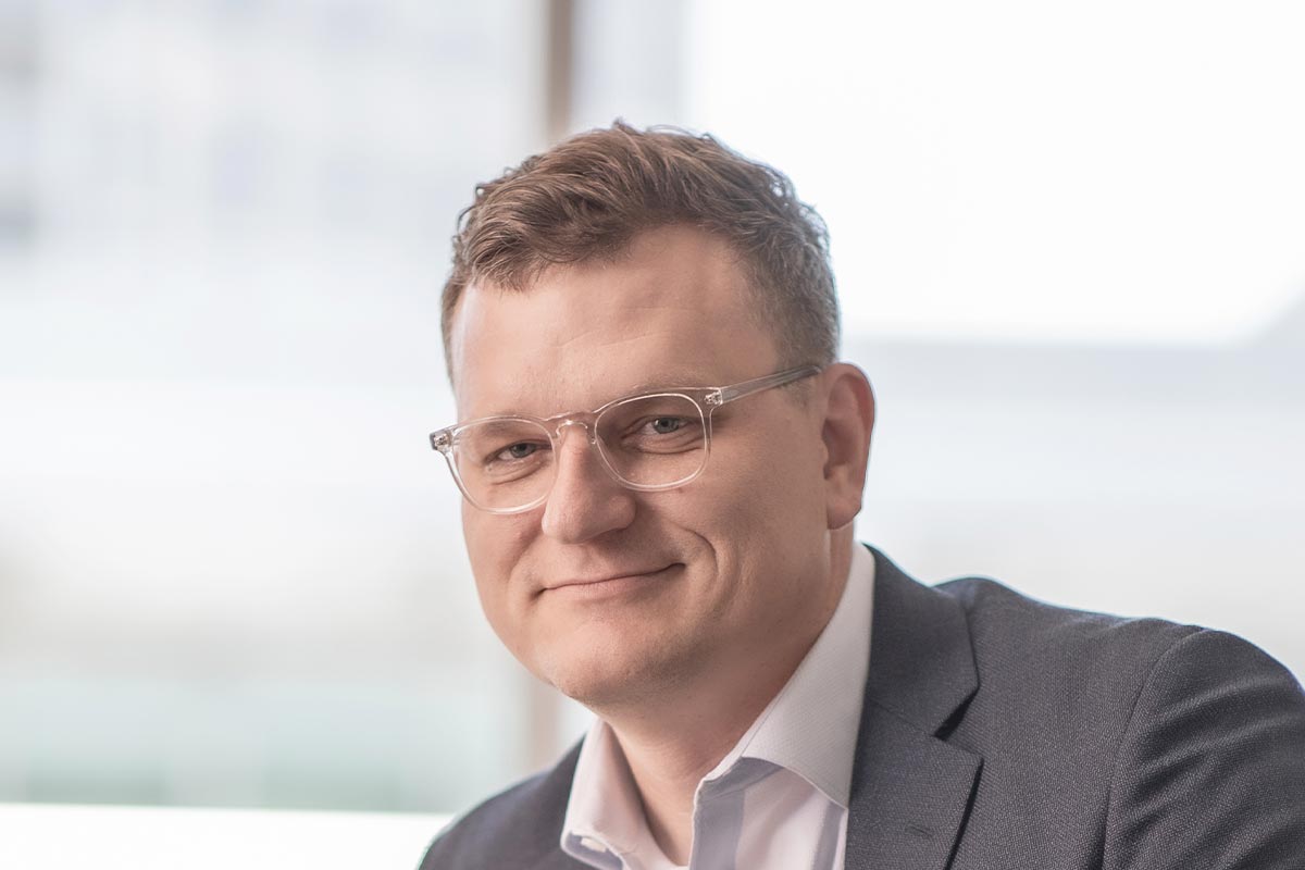 Close-up portrait of John Jacobson wearing clear glasses and a dark blazer, with a softly blurred office background.