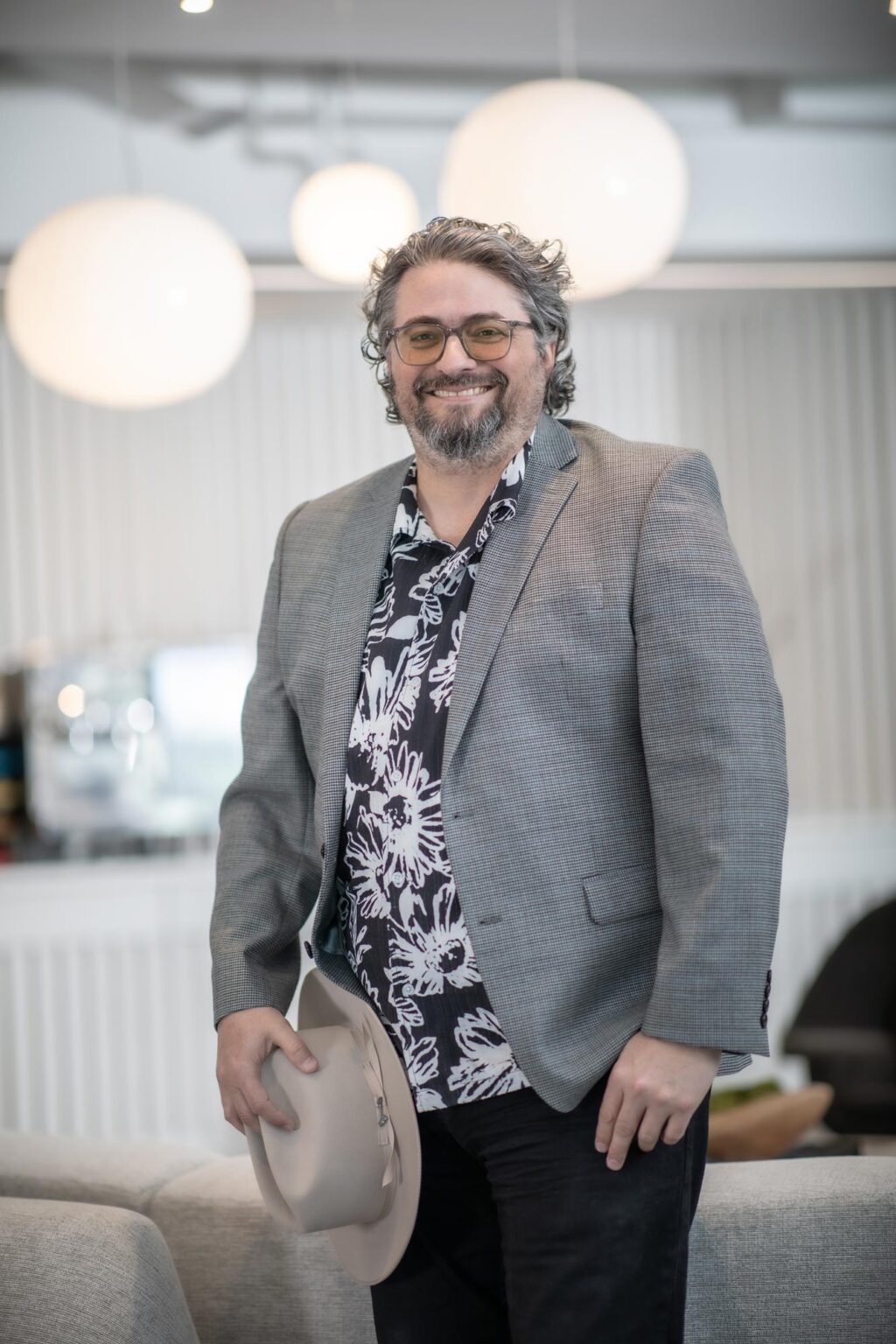 Loren Boynton, Senior Project Leader at GFF, standing and smiling in a contemporary office with globe pendant lights, holding a light-colored hat.